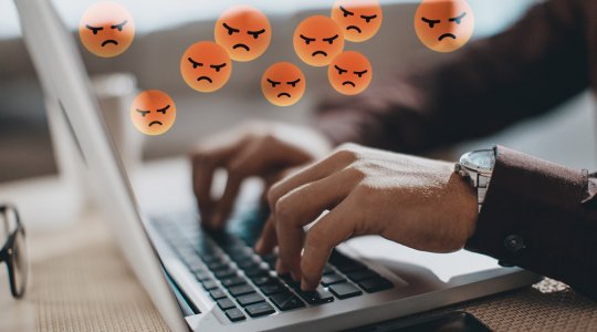 Närbild på händer som skriver på ett tangentbord med arga emojis ovanför som symbol för näthat – Framfot företagsutbildning.