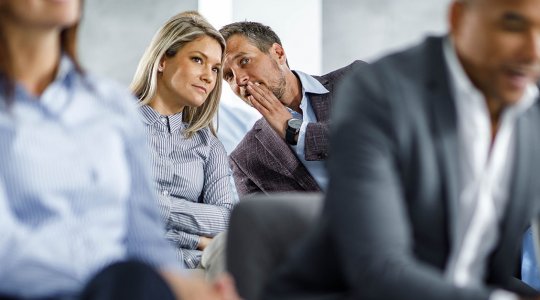 Kvinna och man som viskar till varandra bakom ryggen på kollega